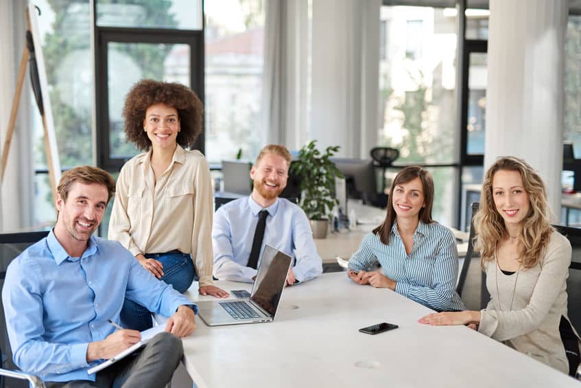 Cinq collègues souriants en réunion dans un bureau moderne avec ordinateurs portables.