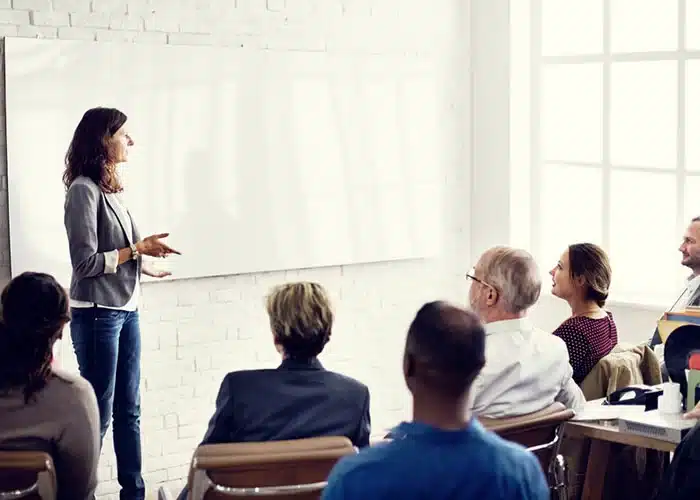 Femme animant une formation devant un groupe attentif dans une salle de réunion lumineuse.